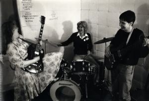 Black and white photo of band rehearsing, Steph with guitar tilted skyward, Angie on drums and Sue on bass guitar, all laughing