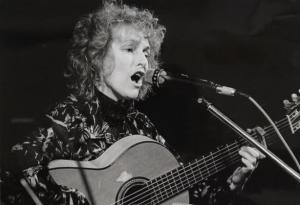 Black and white image of Maria Tolly singing into a microphone, playing an acoustic guitar.