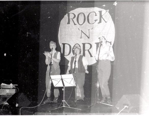 A trio of women in an old black and white photo, onstage with microphones, singing, one signing. A large banner in background saying 'Rock 'n' Doris'.