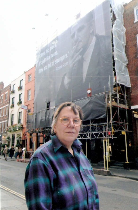 Val Wilmer stands in street in Soho, with a very large banner on the club's outside wall behind her showing founder Ronnie Scott laconically smoking, smiling.